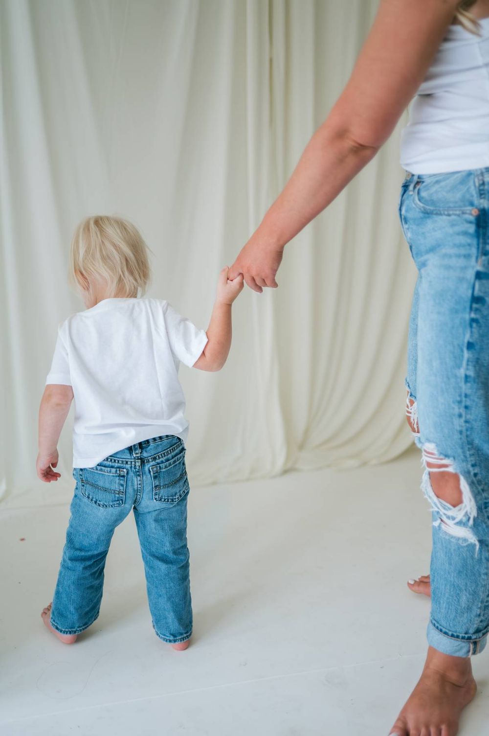 Rear view of a toddler in white shirt and jeans holding mother's hand against a white fabric backdrop Lexington NC