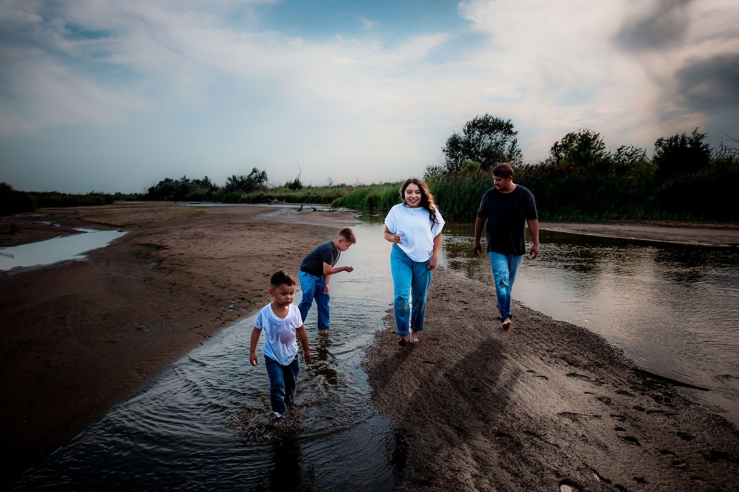 Family walking along a shallow river bank on a cloudy day near lush green trees.