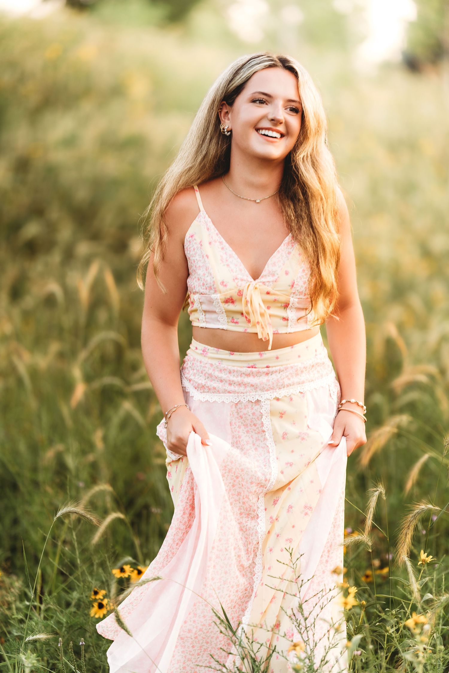Young blonde woman in floral crop top and maxi skirt smiling outdoors in a sunlit meadow.