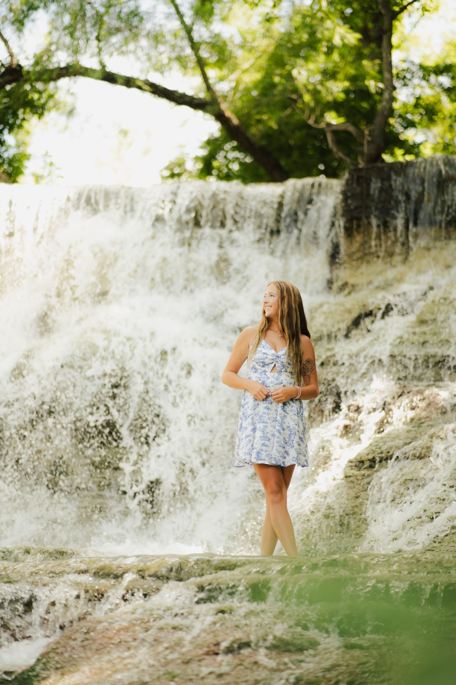 Young woman in blue floral dress standing gracefully in front of a cascading waterfall surrounded by lush green trees.