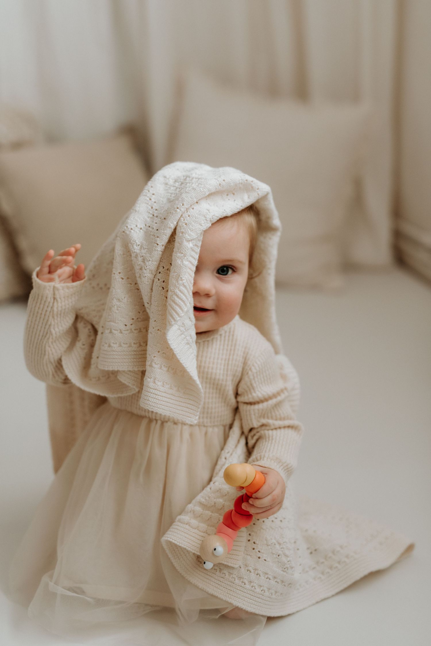 Adorable baby girl in cream knit dress with blanket on head, holding colorful rattle toy, sitting on floor.