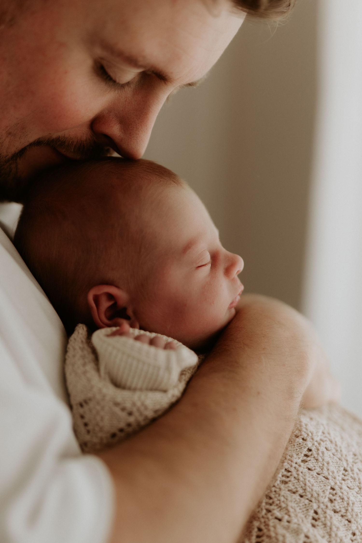 A bearded father tenderly kisses his sleeping newborn baby wrapped in a white knit blanket.
