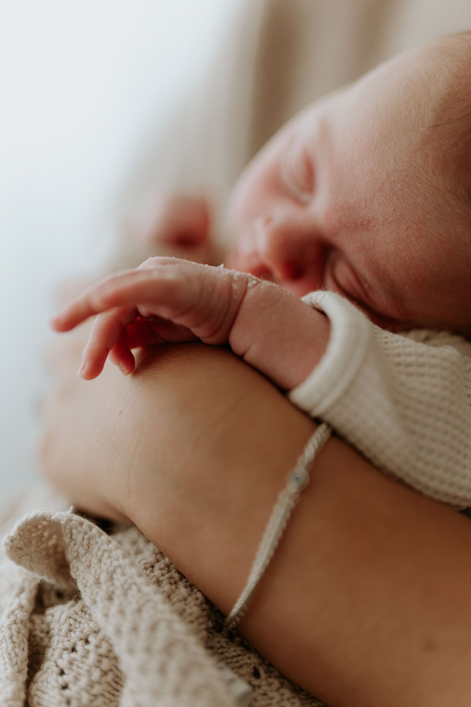 Close-up of a sleeping newborn baby with tiny fingers resting on a parent's arm, wrapped in a soft knit blanket.
