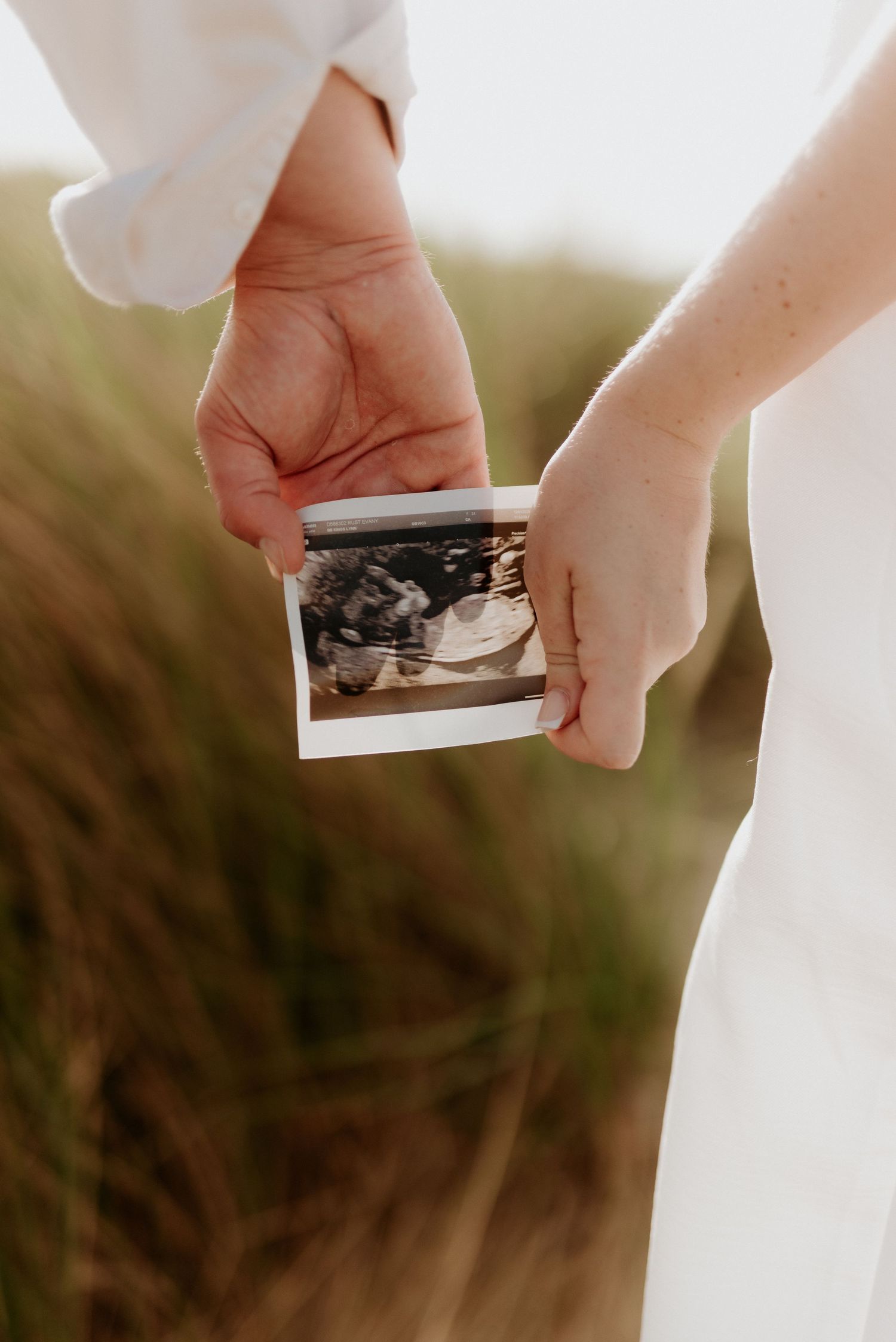 Couple holding a sonogram ultrasound photo outdoors in a grassy field, pregnancy announcement portrait.