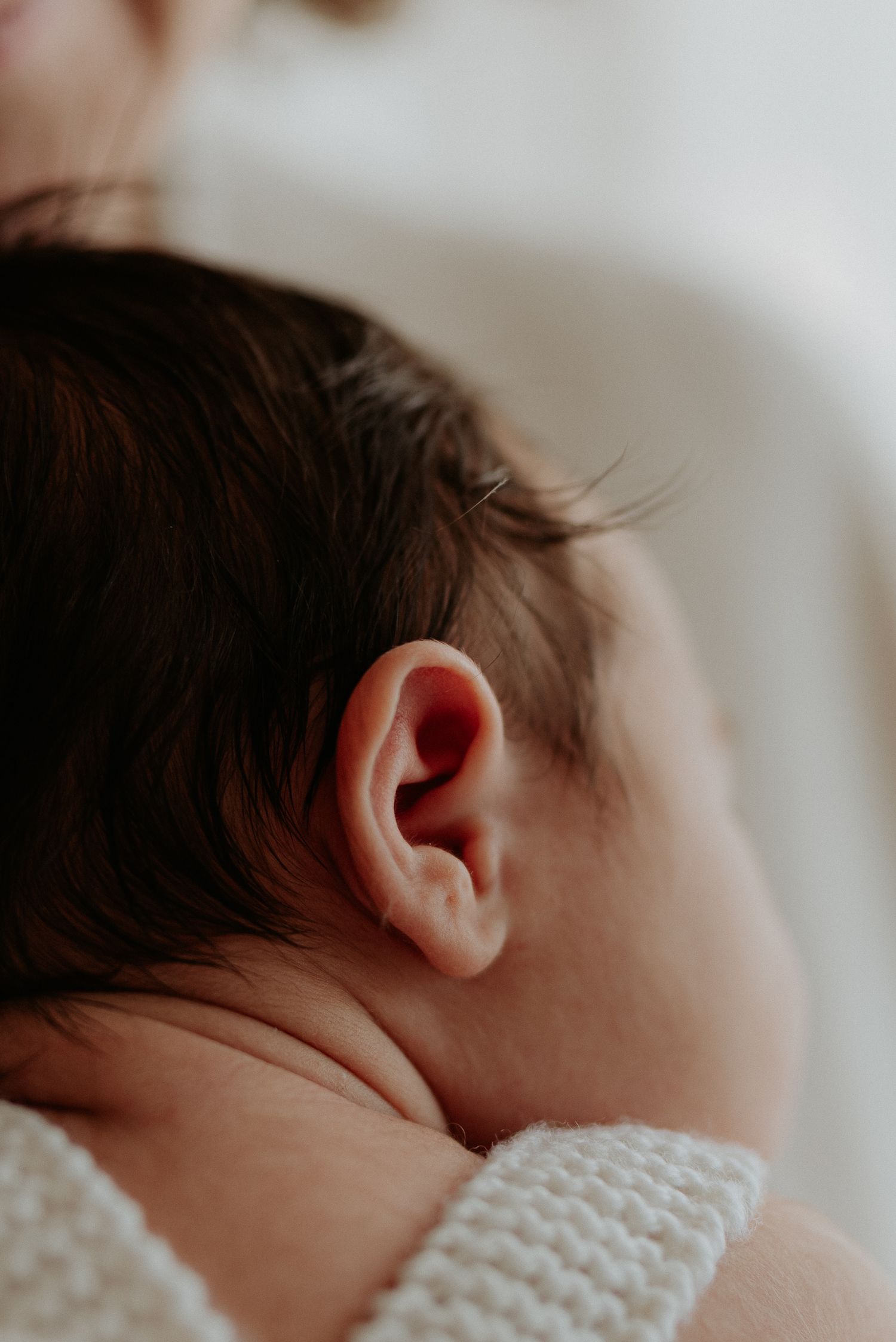 Close-up of a newborn baby's ear with dark hair, wearing a white knit outfit, soft natural light.