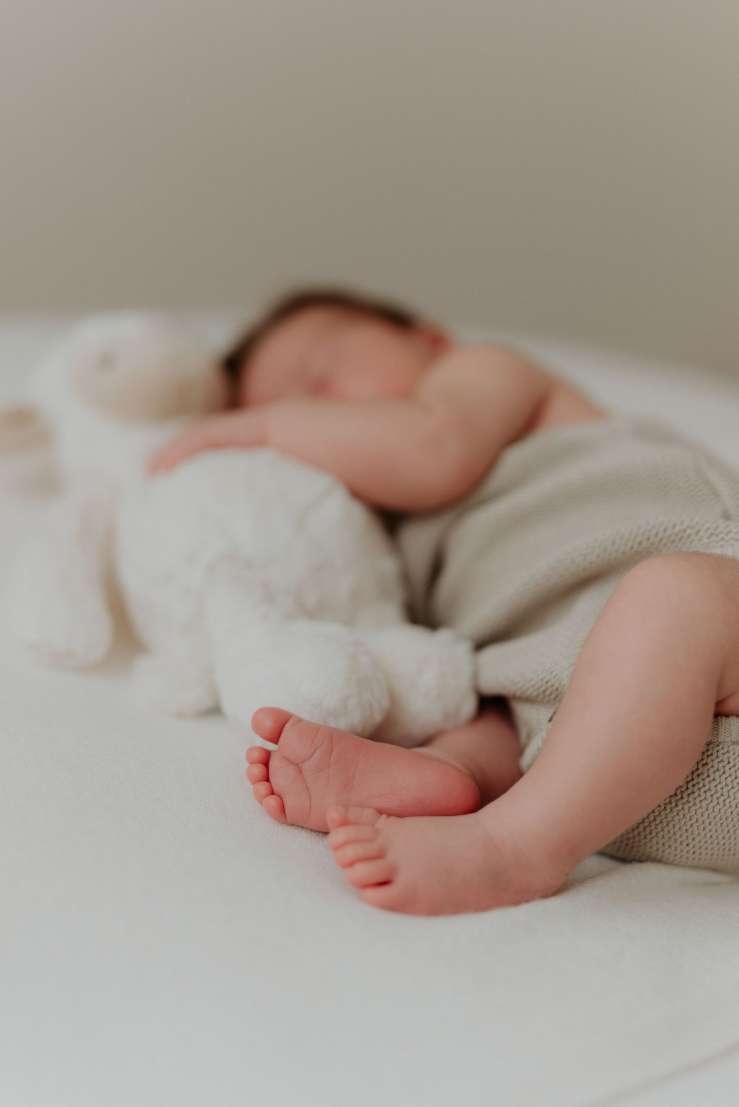 Close-up of a sleeping newborn baby's tiny feet beside a white stuffed bunny toy, wrapped in a soft knit blanket.