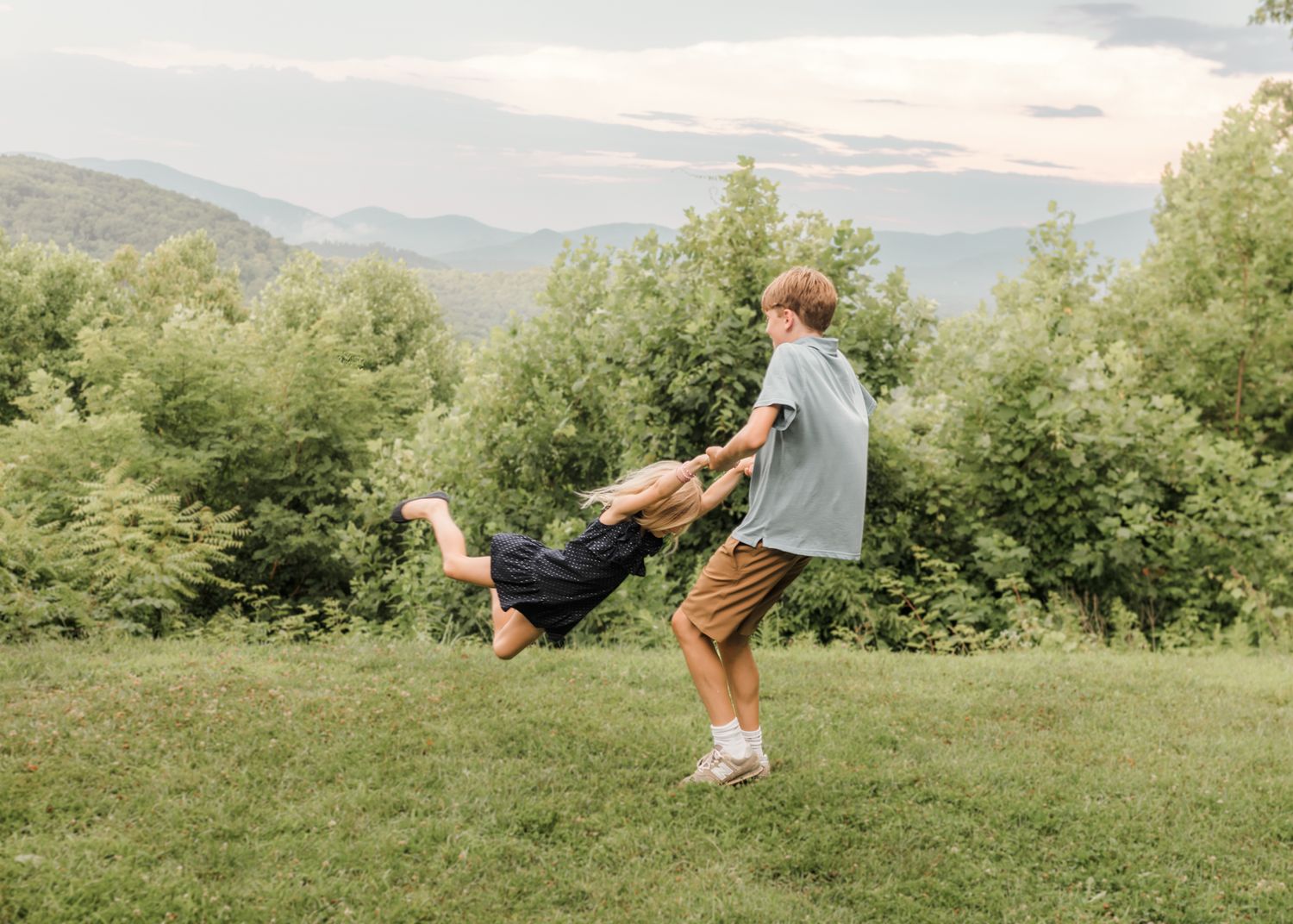 A boy swings a child in a circle on a grassy hill with lush green trees and mountains in the background.