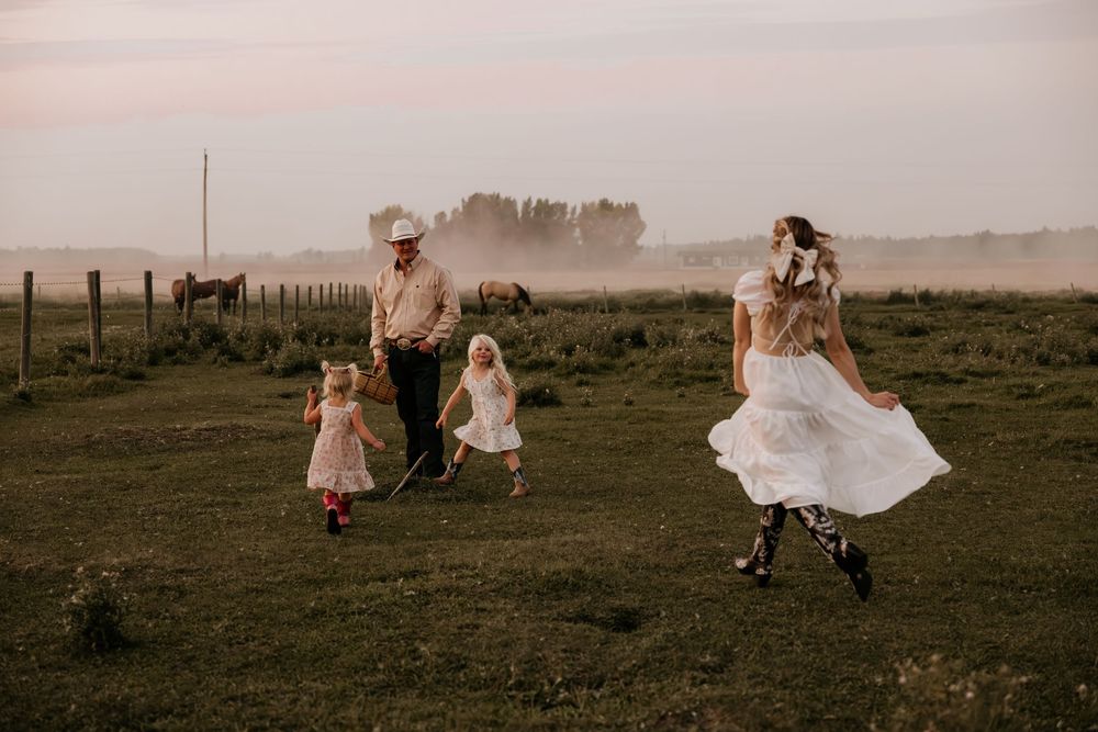 Family in rustic field at sunset, bride in white dress, man in cowboy hat, two young girls playing on green grass with horses.