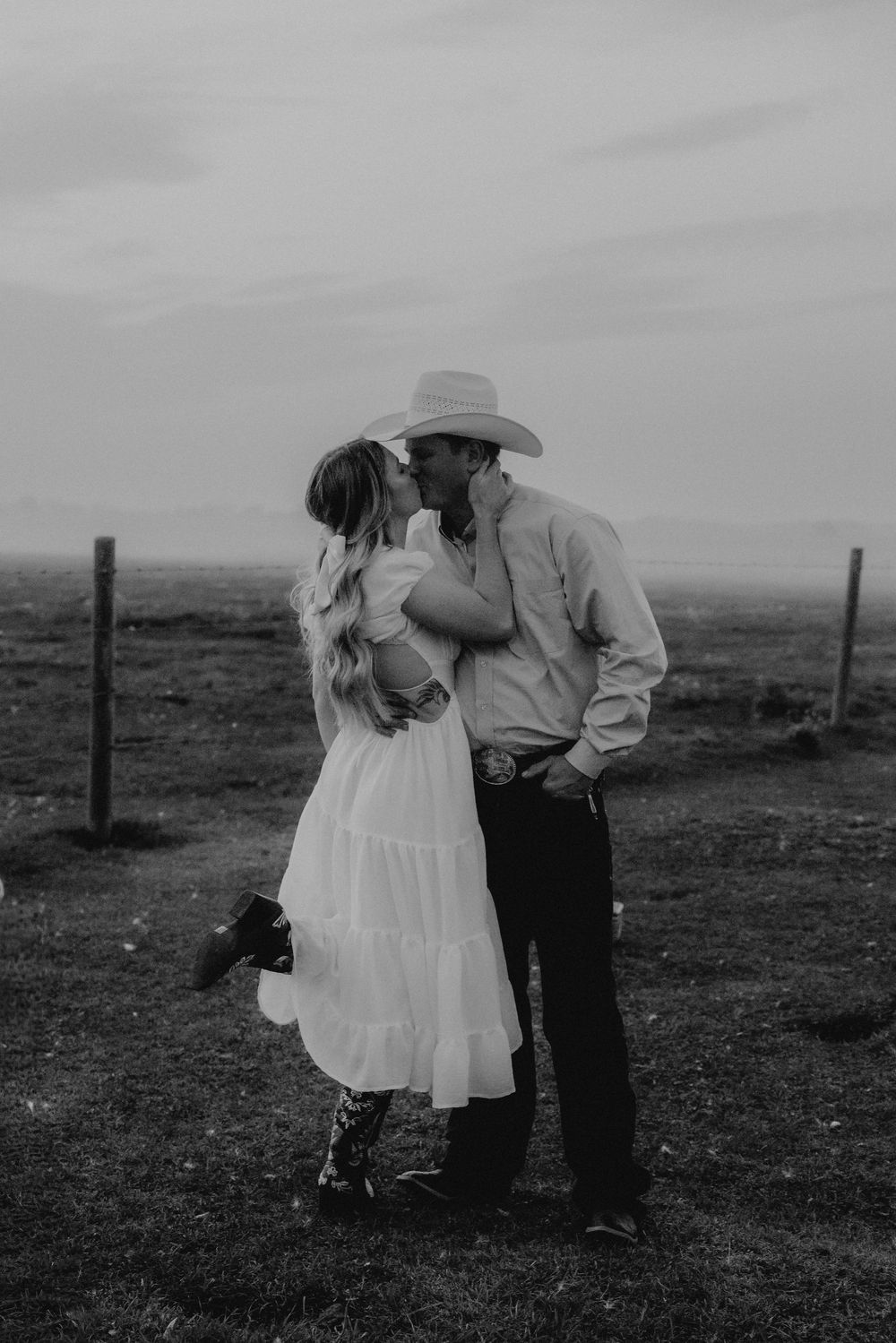Black and white photo of a couple kissing in a field, man wearing cowboy hat, woman in white dress.