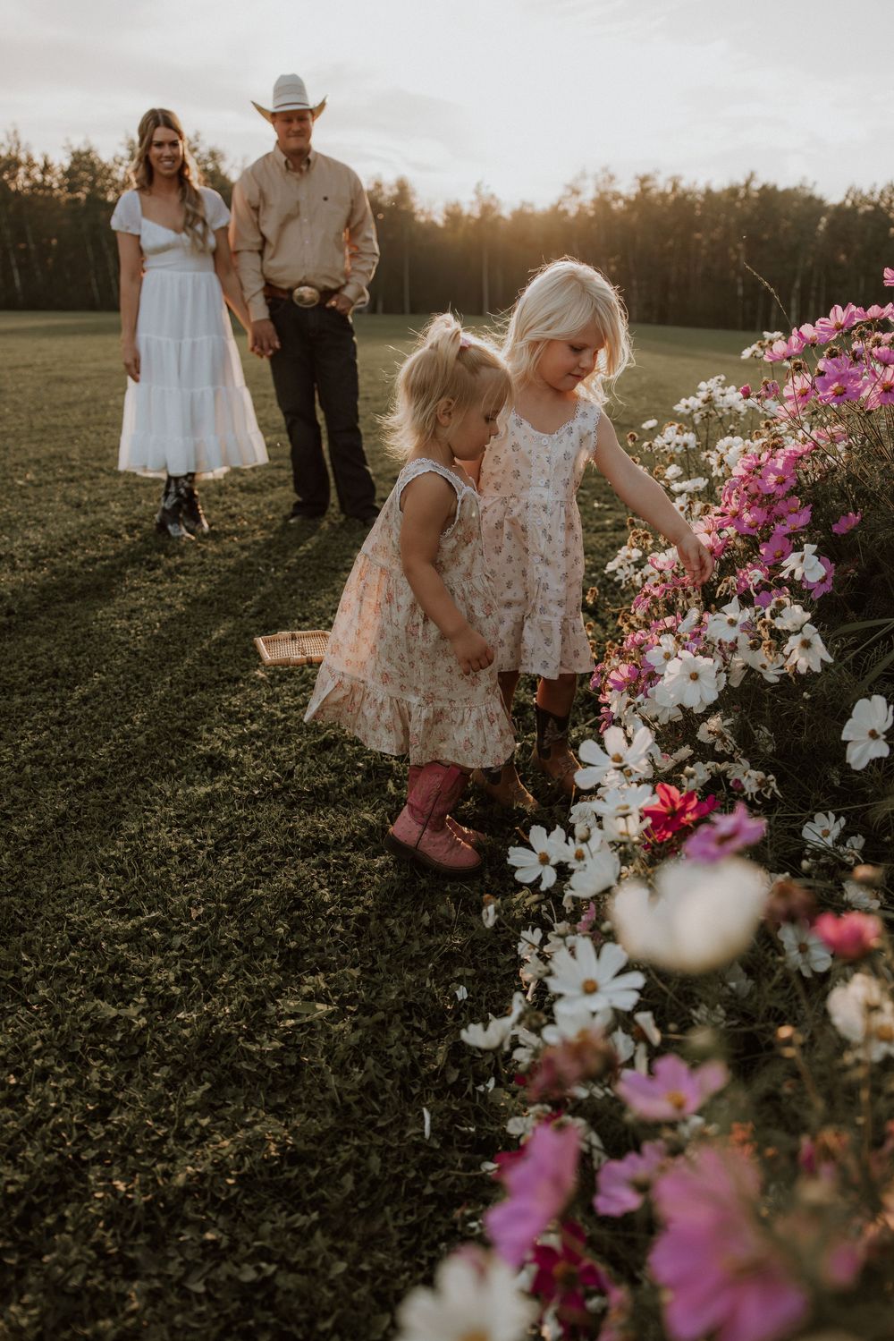Two young girls in floral dresses and pink boots pick flowers at golden hour while parents watch in a field.
