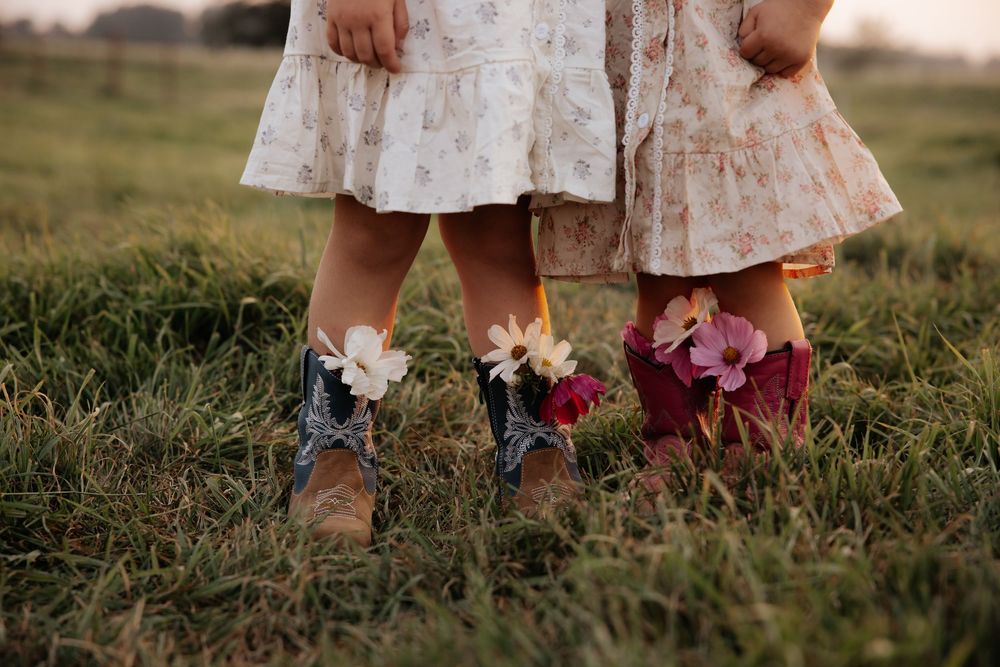 Two girls in floral dresses and cowboy boots stand on grass, boots adorned with colorful wildflowers.