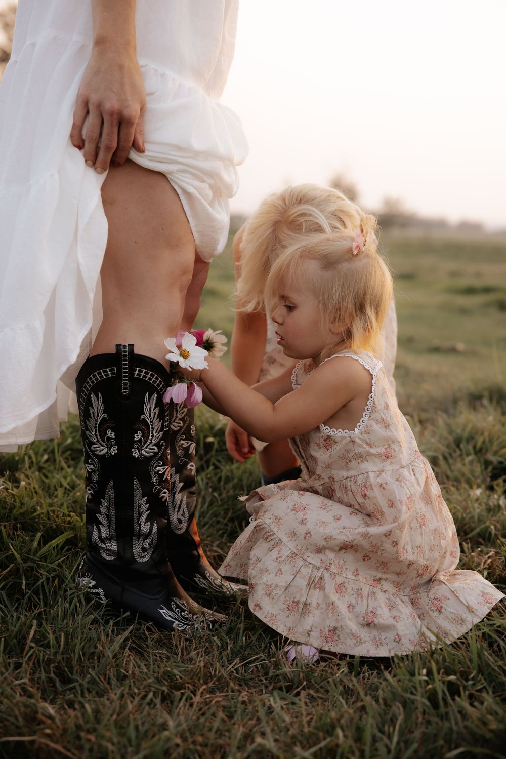 A toddler girl in a floral dress places flowers into a woman's black cowboy boots while outdoors in a grassy field.