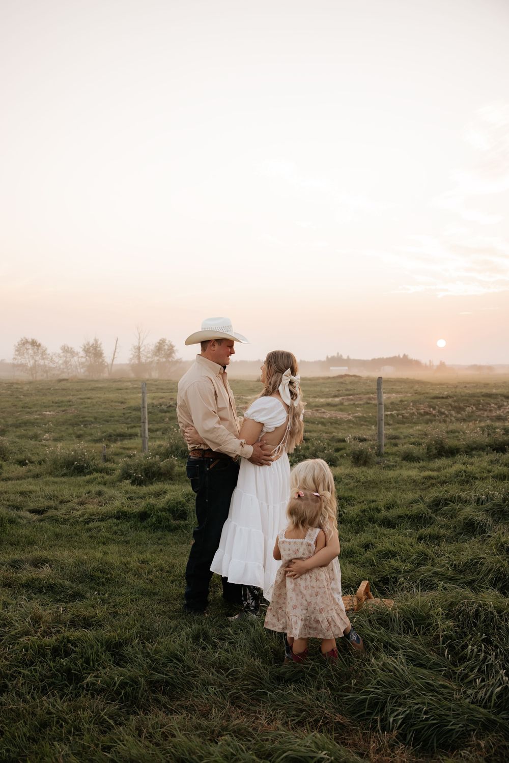 A family of three stands in a lush green field at golden sunset, with a man in a cowboy hat embracing a woman in a white dress.