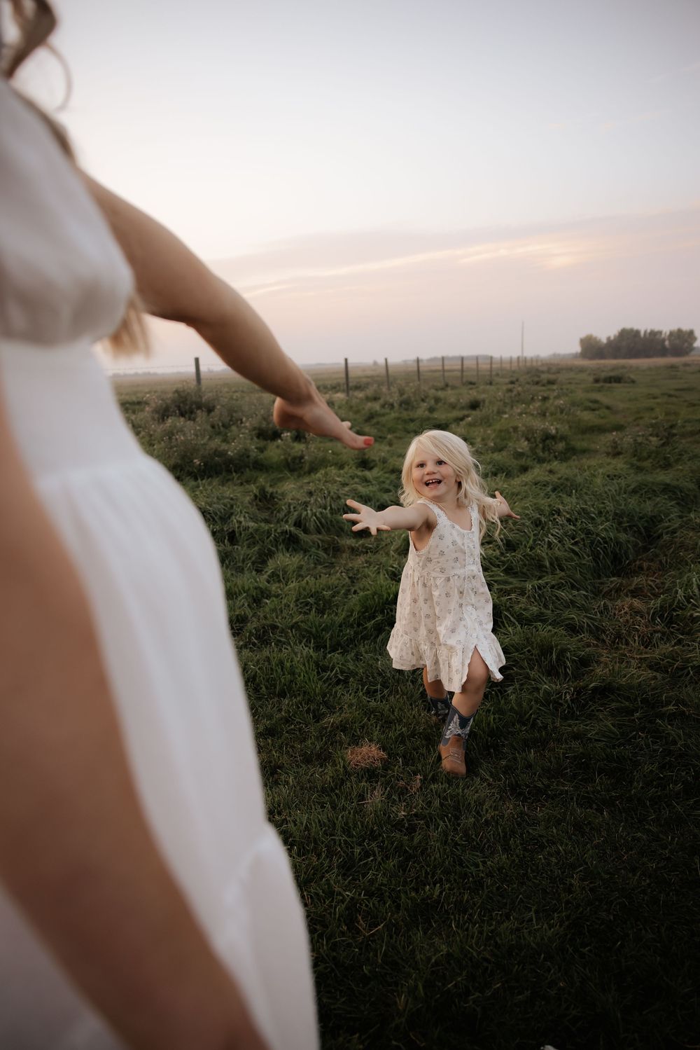 A joyful blonde toddler in a white floral dress runs toward her mother across a lush green field at sunset.