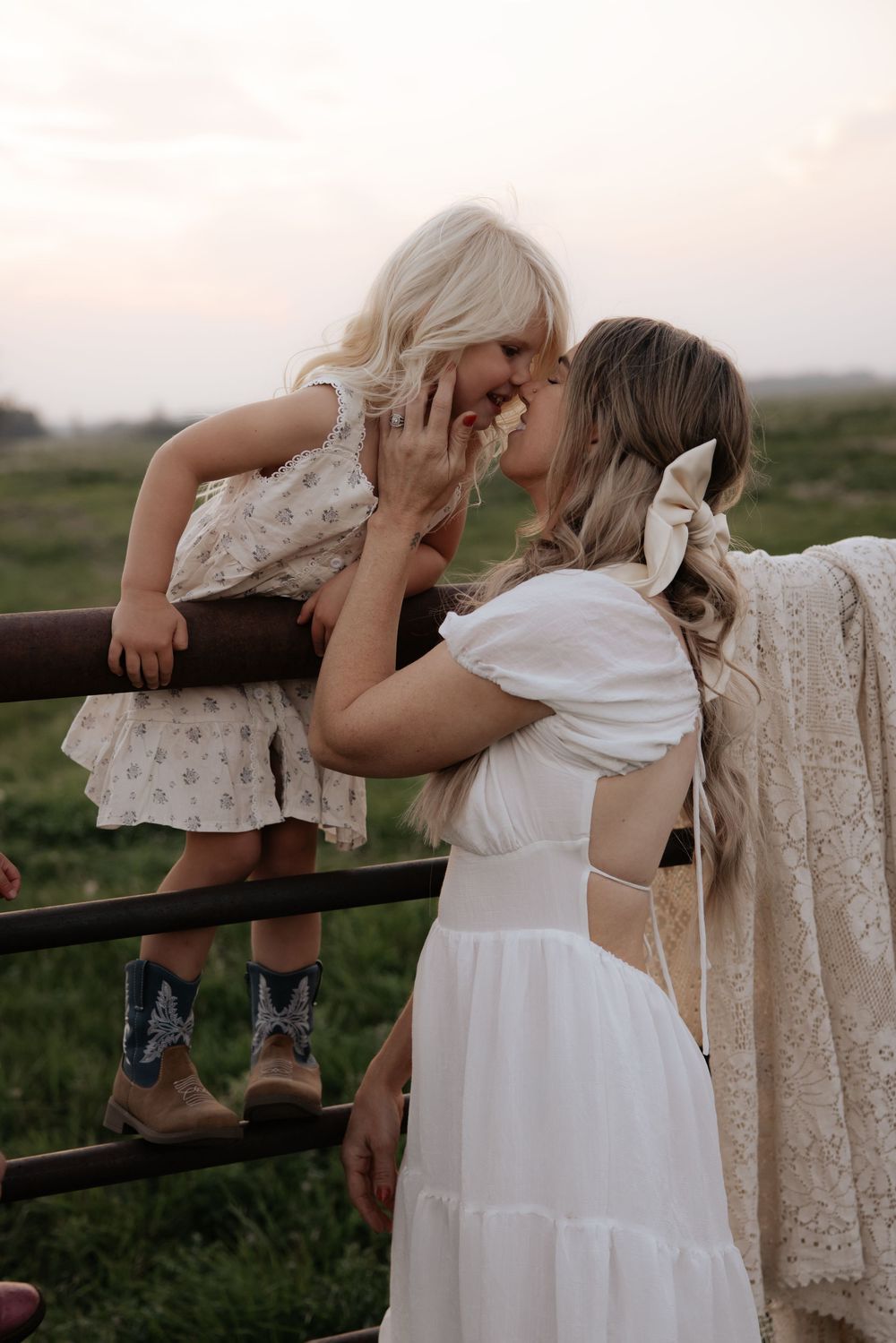 A woman in a white dress shares a tender nose-to-nose moment with a young blonde girl sitting on a wooden fence outdoors.