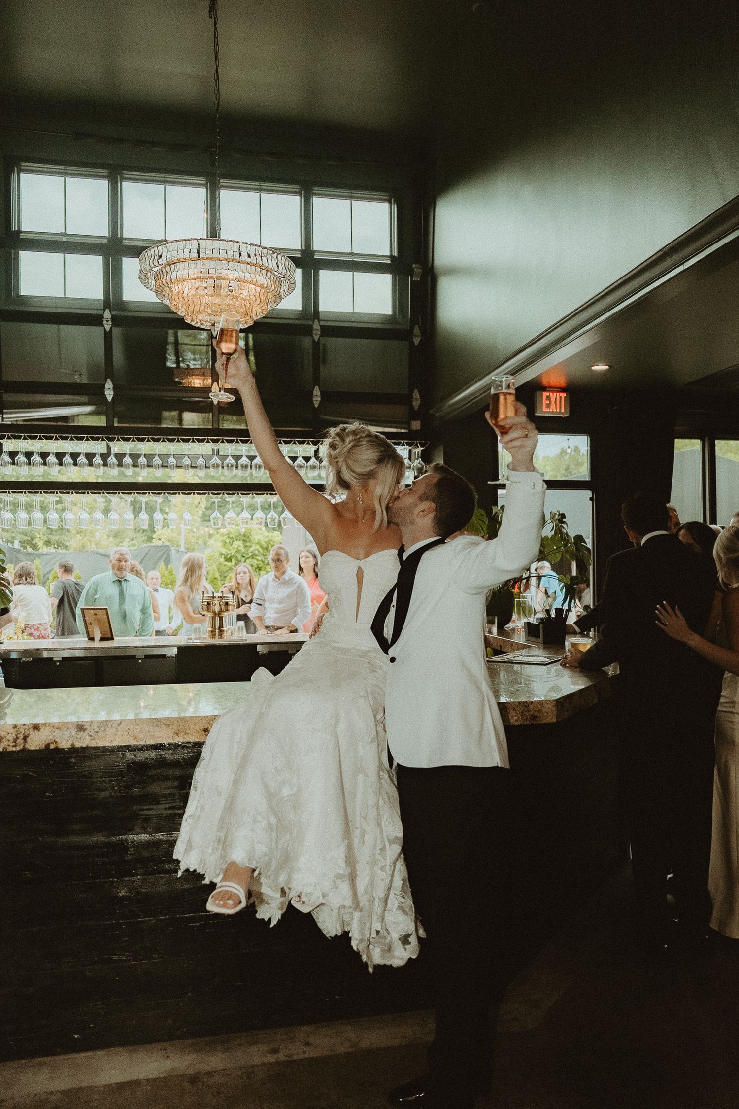 Newlyweds kiss and raise glasses at wedding reception, bride in white gown, groom in white suit, elegant chandelier above.