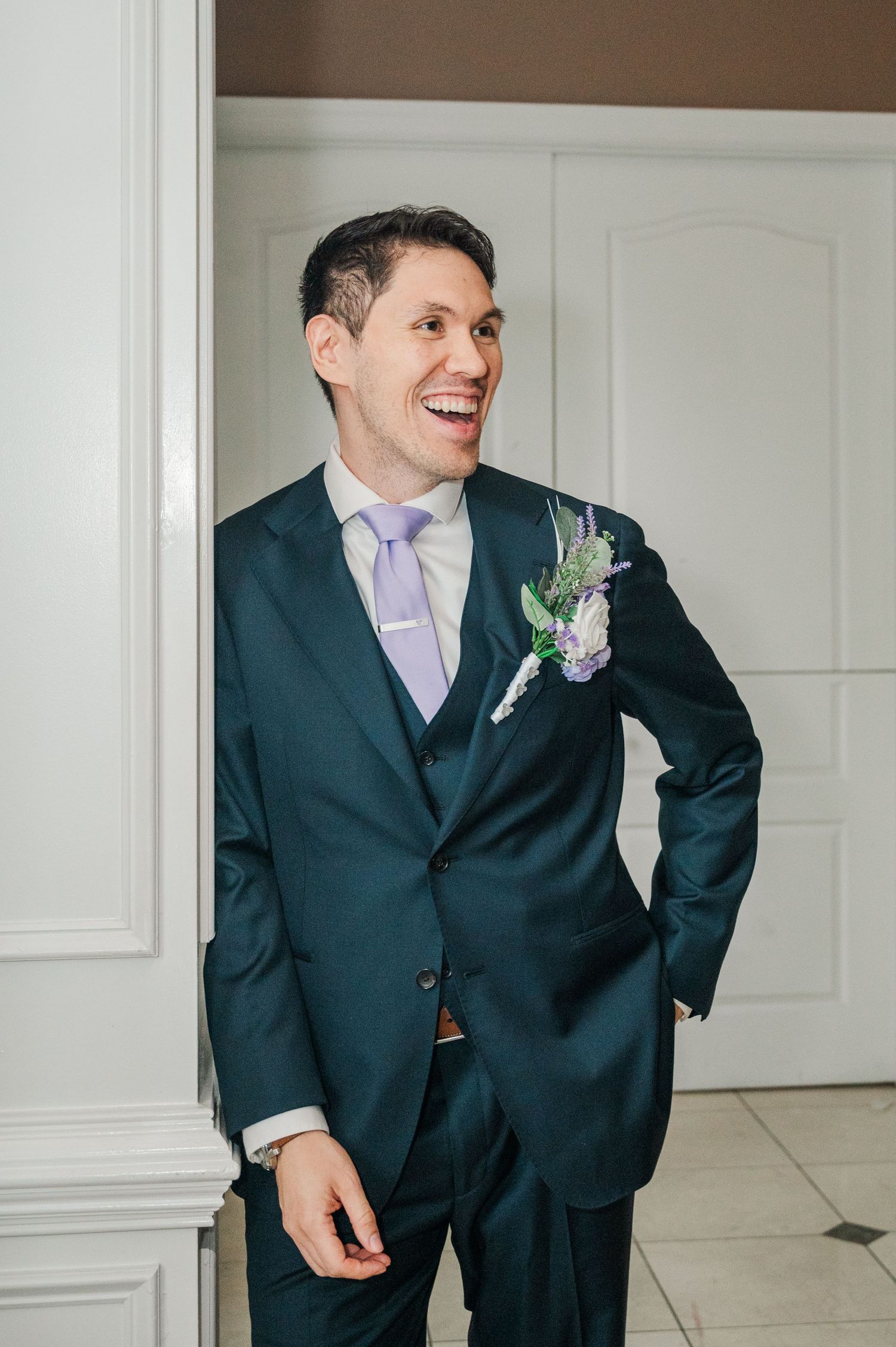 Smiling groom in dark teal suit with lavender tie and floral boutonniere laughing joyfully on his wedding day.