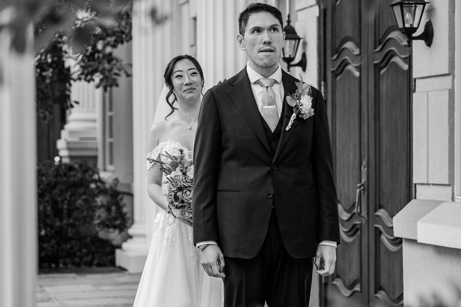 Black and white photo of a bride and groom standing together outside a venue on their wedding day.