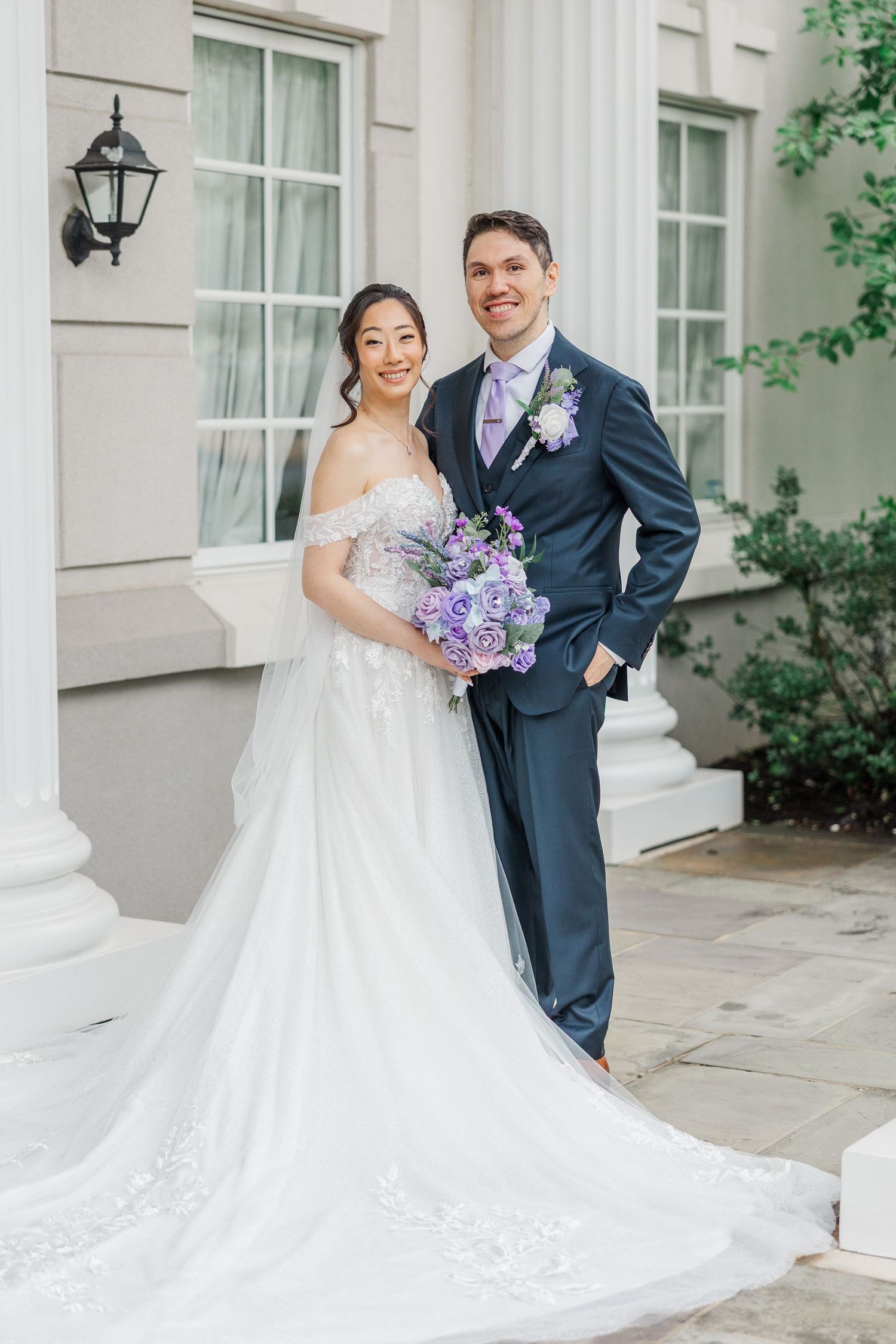 Bride in off-shoulder gown holds purple bouquet beside groom in navy suit outside elegant white building.