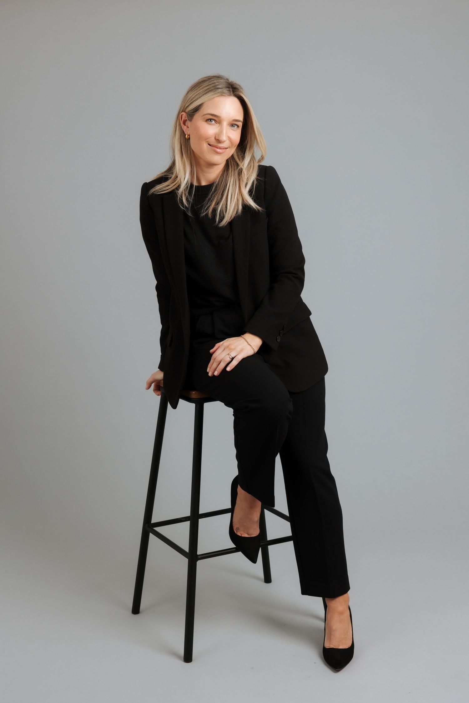 Woman in black blazer and trousers sitting on a stool against a gray background, smiling confidently.