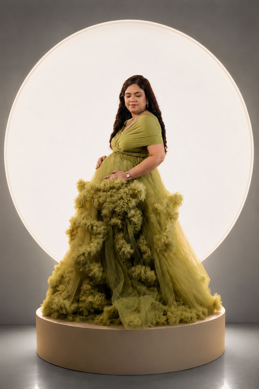 Pregnant woman in olive green ruffle maternity gown poses gracefully in front of a glowing circular light backdrop.