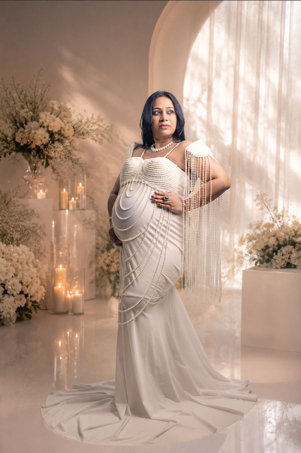 Pregnant woman in elegant white beaded gown poses gracefully among candles and white florals in a soft, luminous studio setting.
