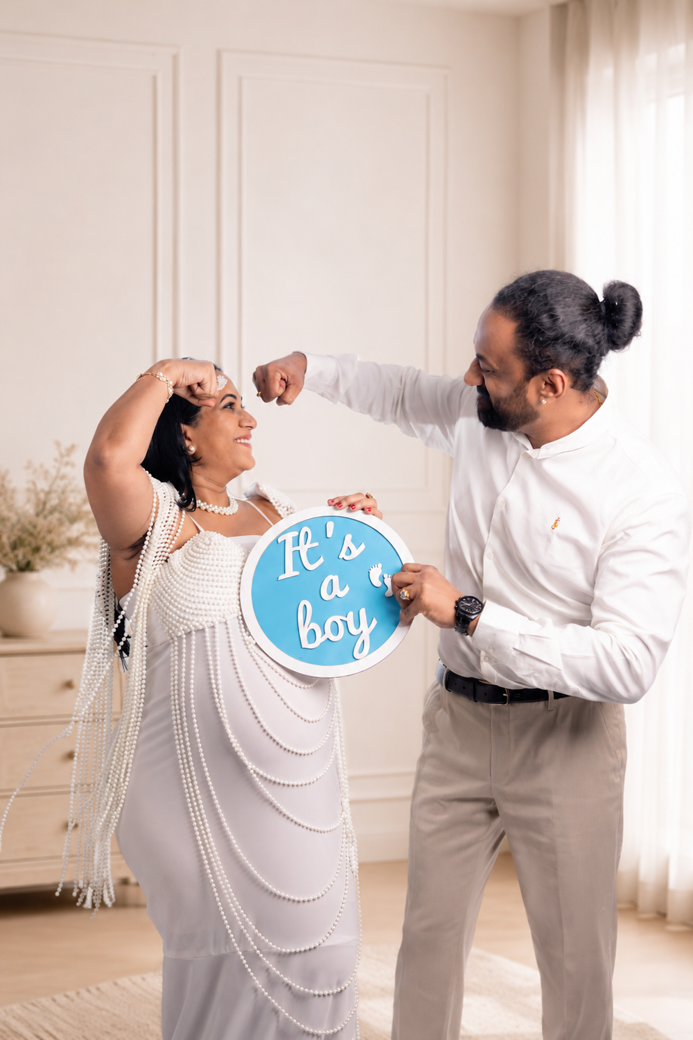 Couple celebrating gender reveal holding blue It's a Boy sign, pregnant woman in white pearl dress, indoor setting.