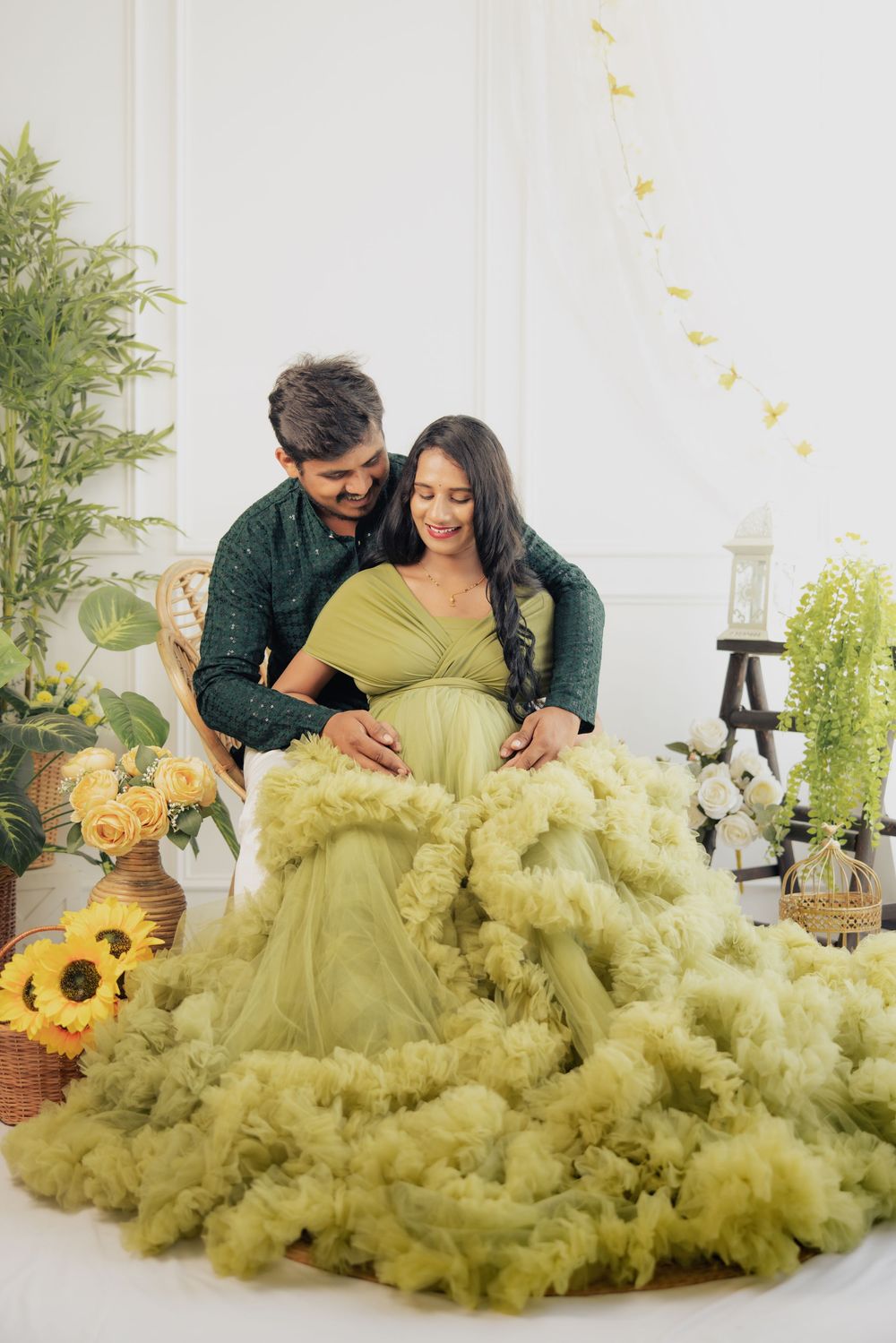 Couple in maternity photoshoot, woman in green tulle gown, man in green shirt, surrounded by flowers and plants.