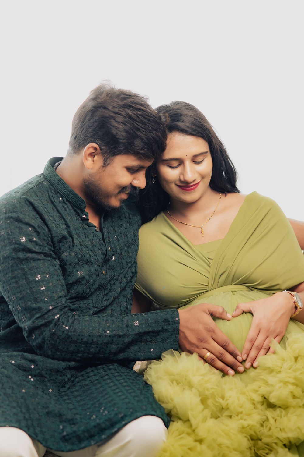 Indian couple in green attire posing for maternity photoshoot, making heart shape on pregnant belly.