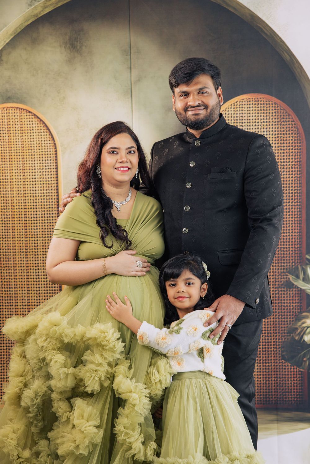 Pregnant Indian woman in olive green gown poses with husband in black sherwani and young daughter for family maternity photo.