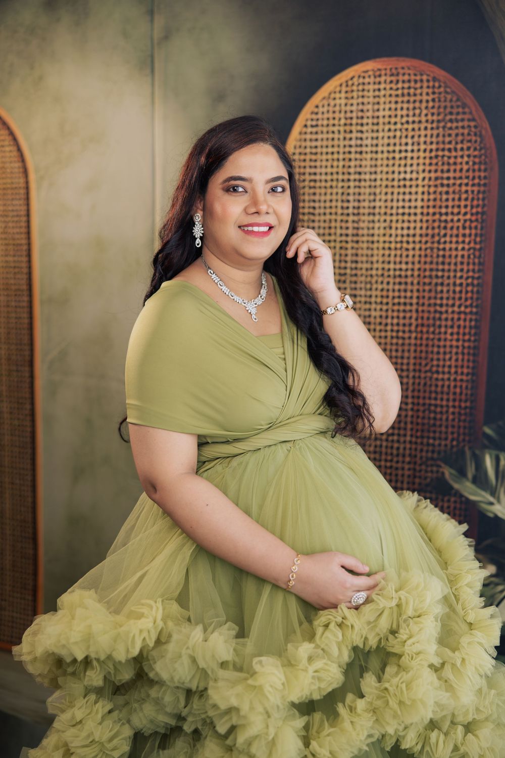 Pregnant woman in olive green ruffled gown with diamond jewelry smiles during maternity photoshoot.
