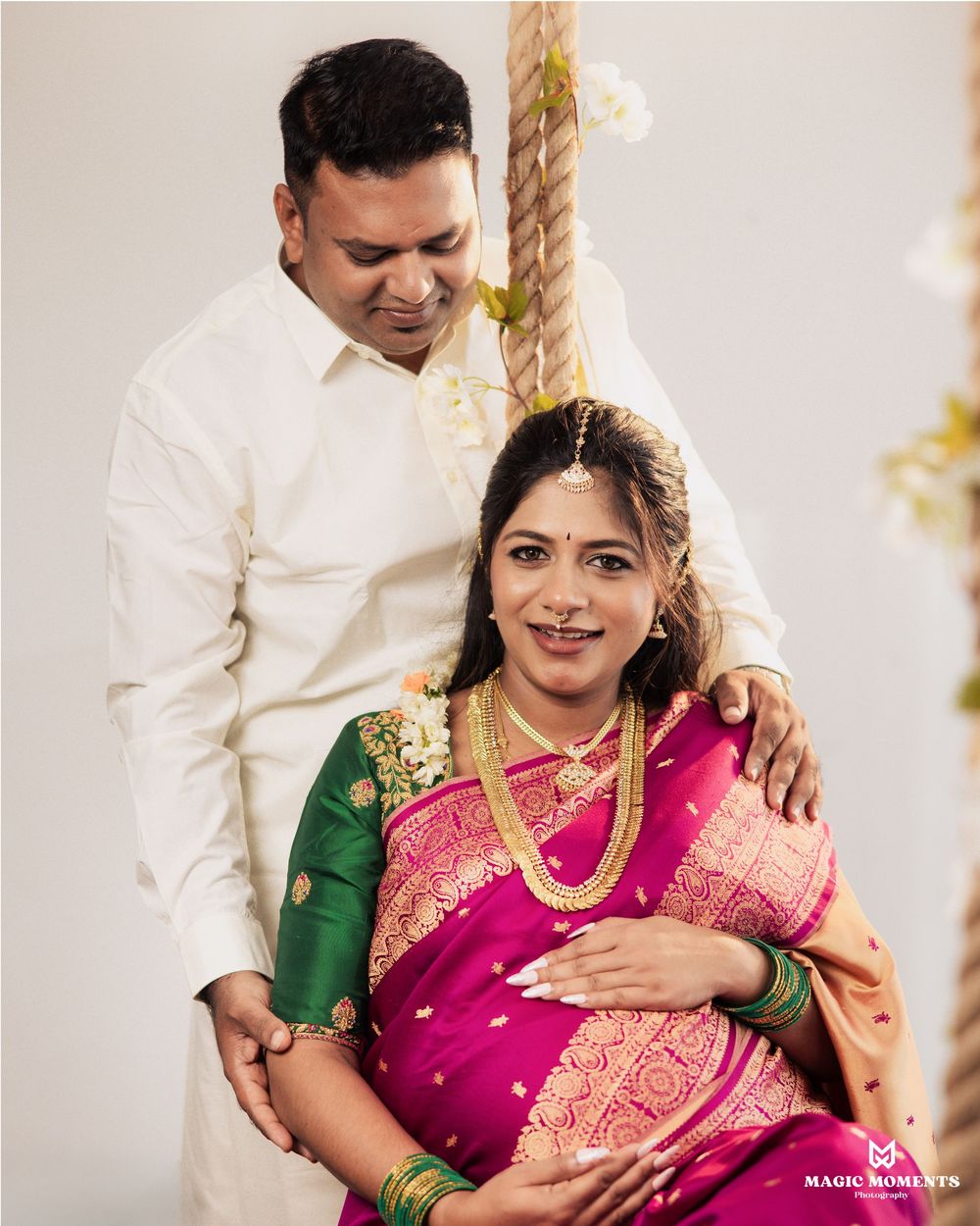 Indian couple in traditional wedding attire, woman in pink saree with gold jewelry, man in white shirt, posing together.