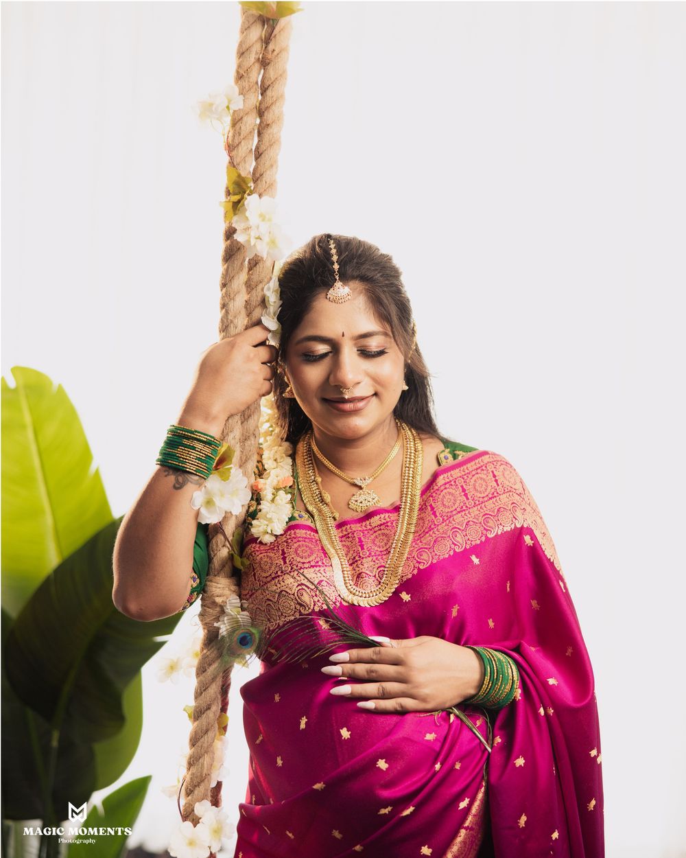 Pregnant woman in pink silk saree with gold jewelry holds a rope swing adorned with white flowers.