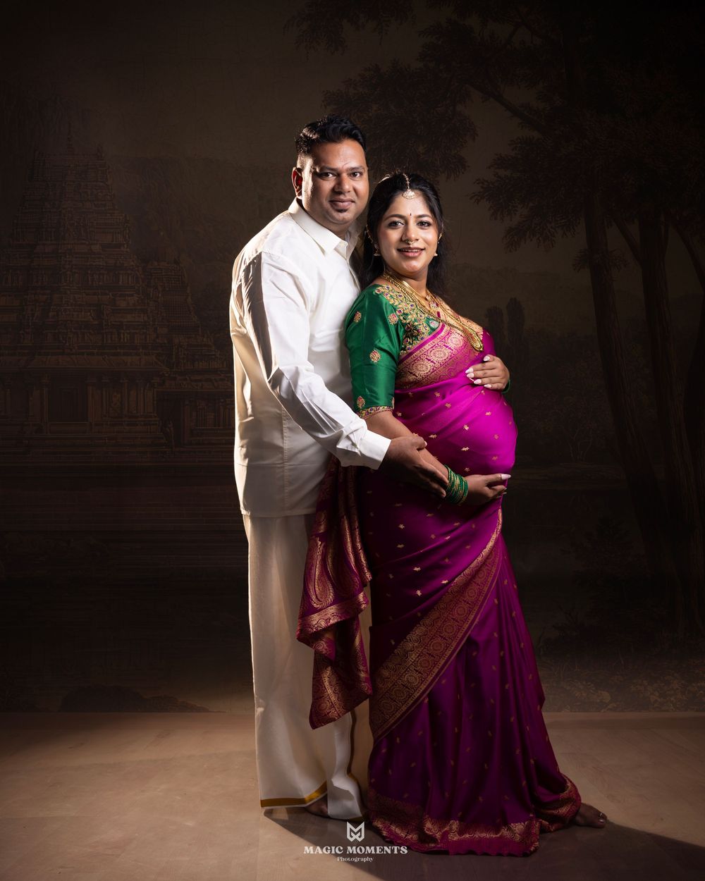 Couple in traditional Indian attire poses for maternity photo, woman in purple saree, man in white kurta, studio backdrop.