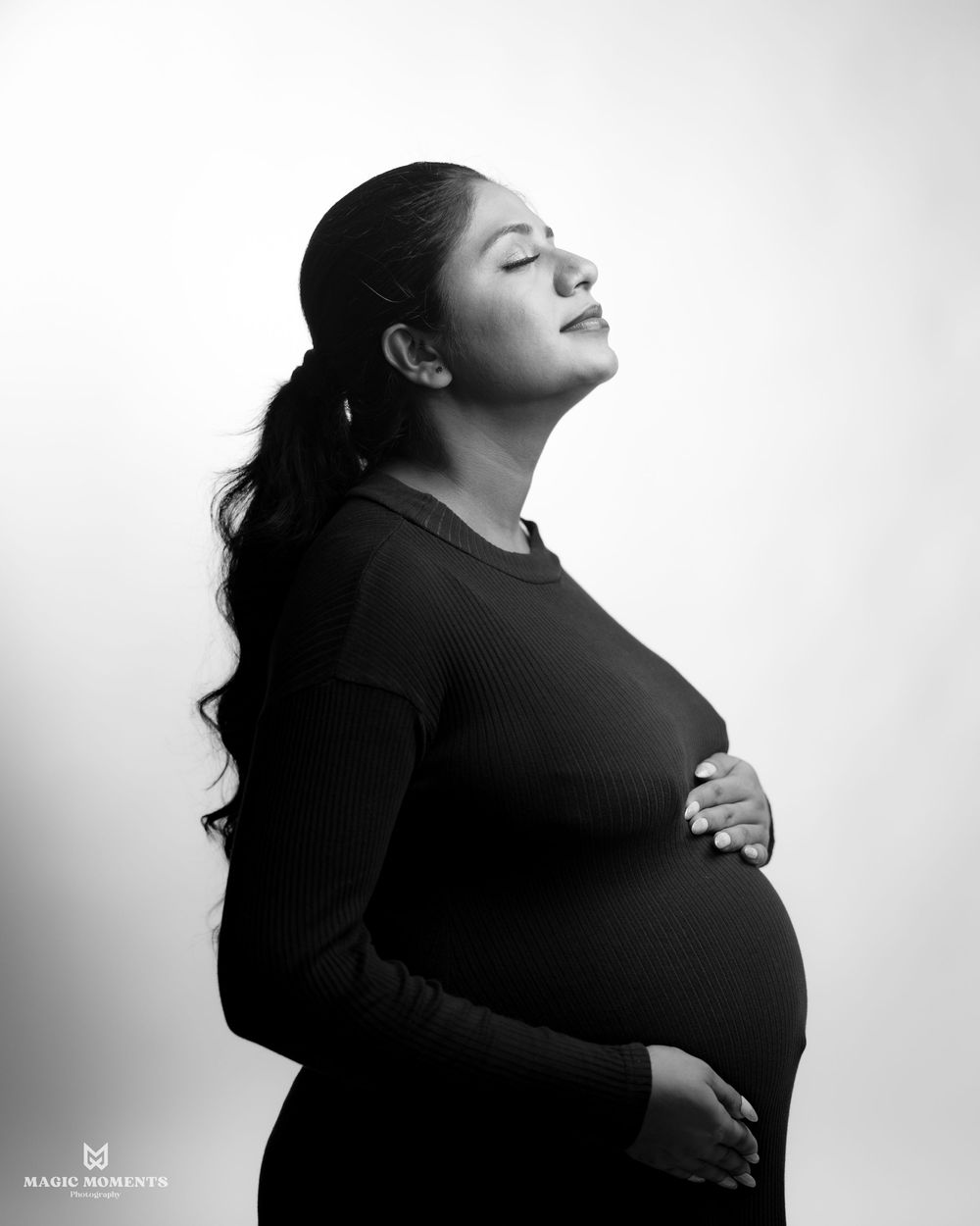 Black and white maternity photo of a pregnant woman in a black dress cradling her baby bump with eyes closed.