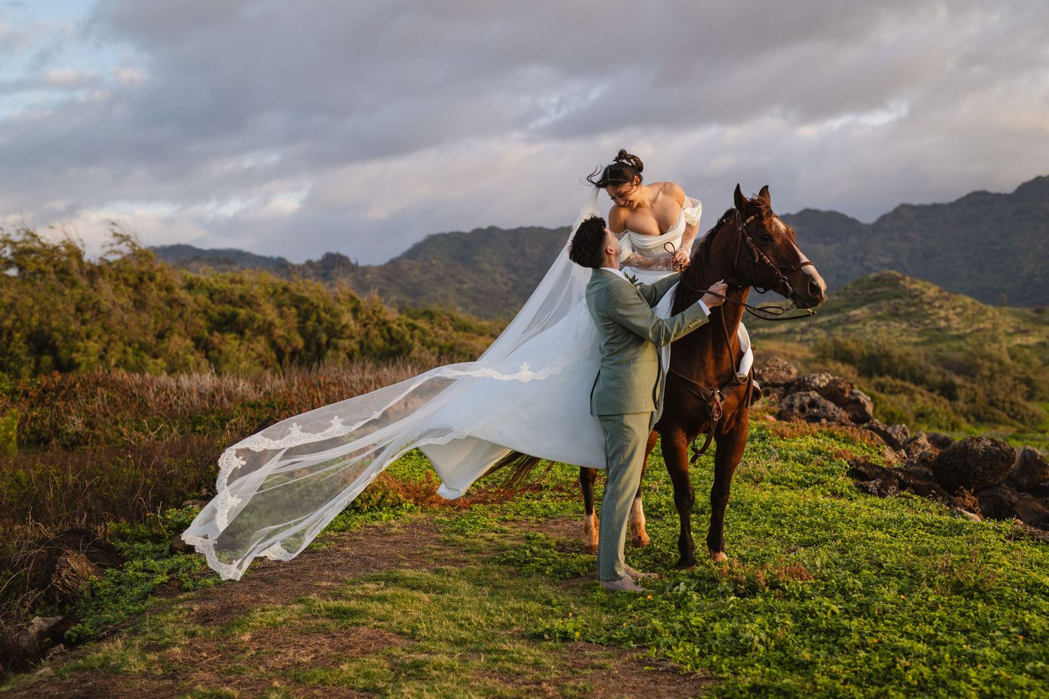 Groom lifts bride in flowing gown beside brown horse on lush green Hawaiian hillside.