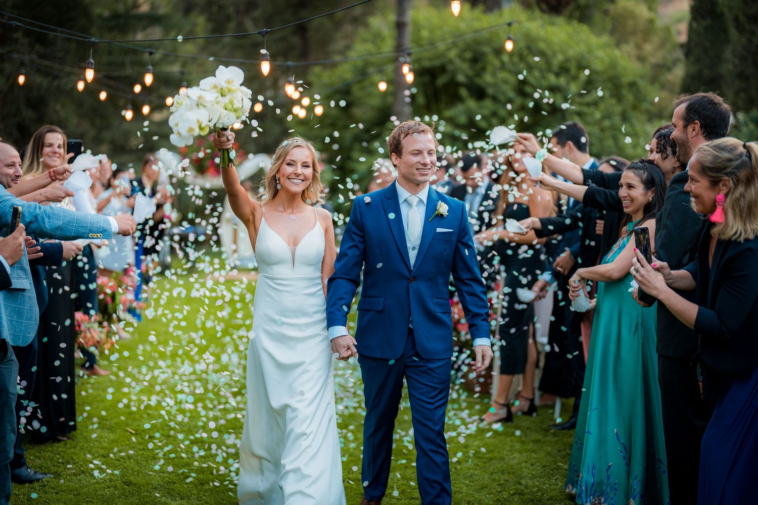 Bride and groom walk through confetti shower, guests cheering, outdoor wedding ceremony, string lights, bride holding bouquet.