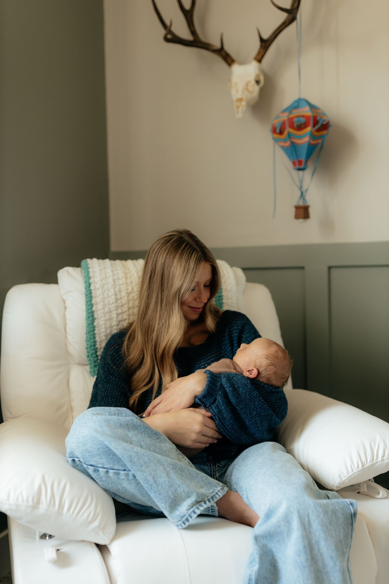 Mother cradling newborn baby in white nursery chair with antler decor and colorful hot air balloon mobile.