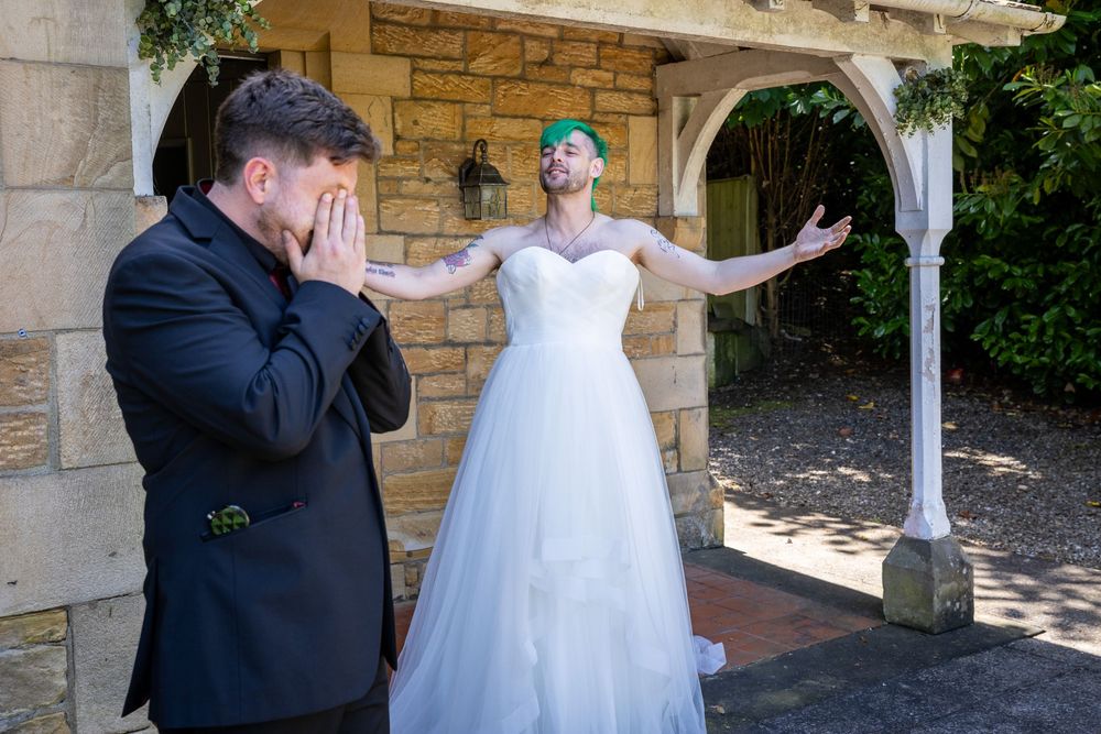 Emotional groom covers his face as bride in white gown stands with arms outstretched during wedding first look.