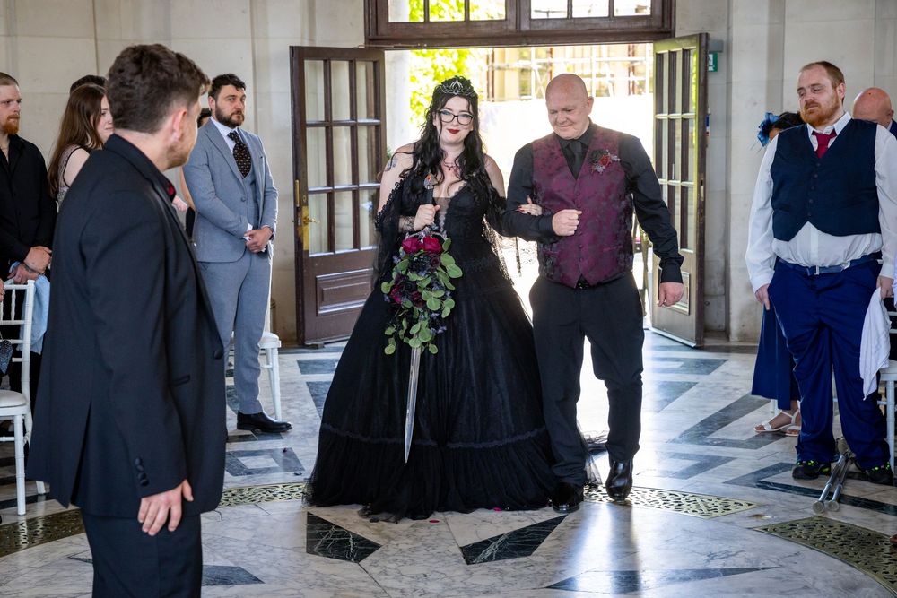 Gothic bride in black wedding dress walks down aisle with escort holding red bouquet at elegant venue.