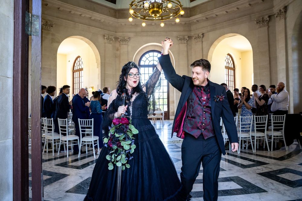 Gothic wedding couple celebrating in elegant hall, bride in black gown with red bouquet, groom in burgundy suit.