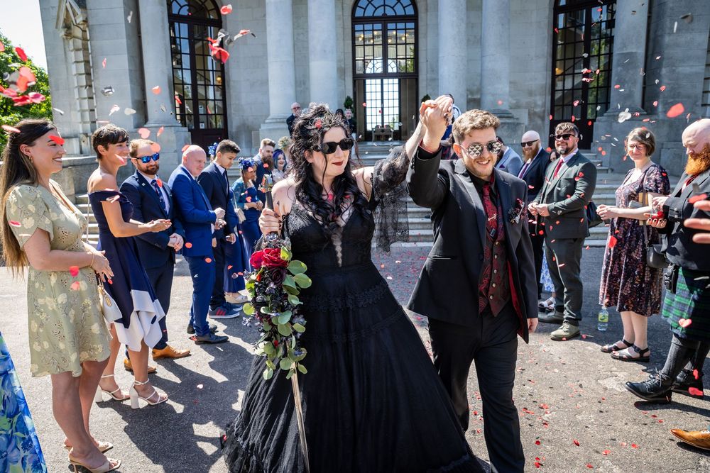 Couple in black gothic wedding attire celebrating outside a grand building with guests throwing confetti.