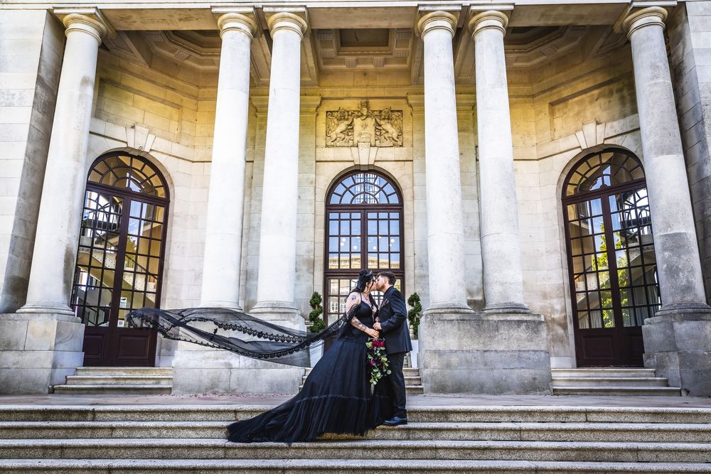Couple in elegant black wedding attire embracing on grand stone steps with tall columns at a classic European venue.