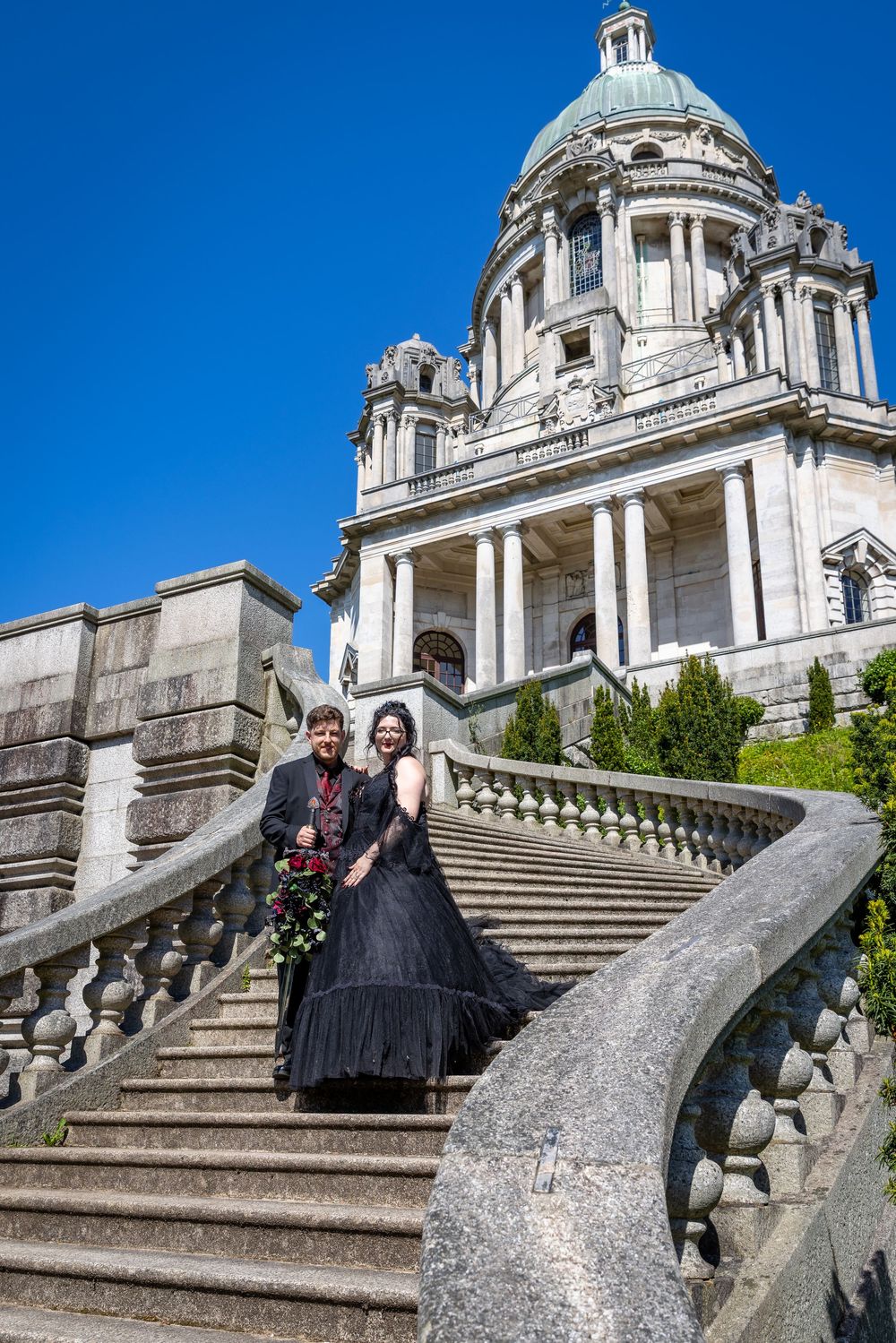 Couple in black wedding attire posing on grand stone steps of a neoclassical building under blue sky.