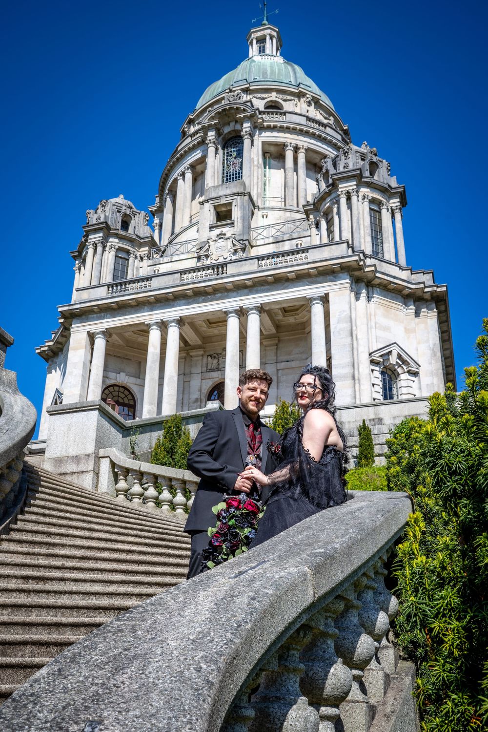 Couple in gothic wedding attire posing on stone steps before a grand neoclassical domed building under a clear blue sky.