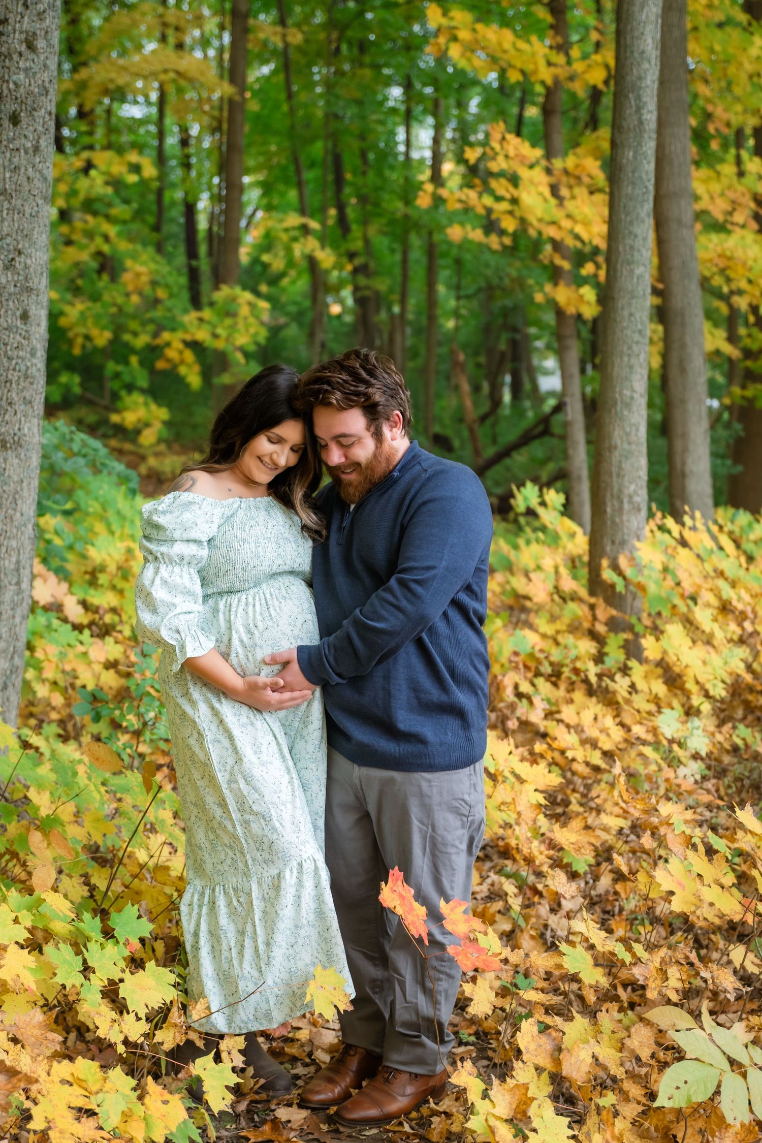 Couple sharing a tender moment in autumn forest, woman in floral dress cradling pregnant belly, man in blue sweater.