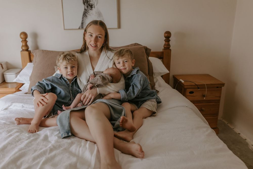A smiling mother in a white robe sits on a bed with her two young children, all wearing robes in a cozy bedroom.
