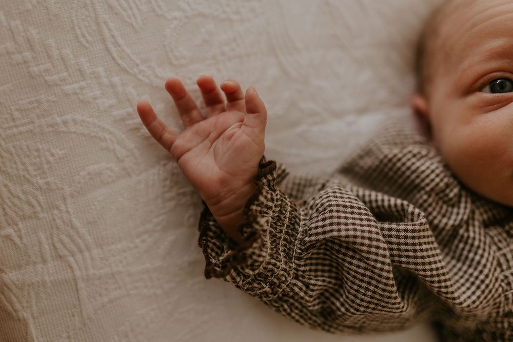 Close-up of a newborn baby's tiny hand resting on a white blanket, wearing a brown gingham outfit.