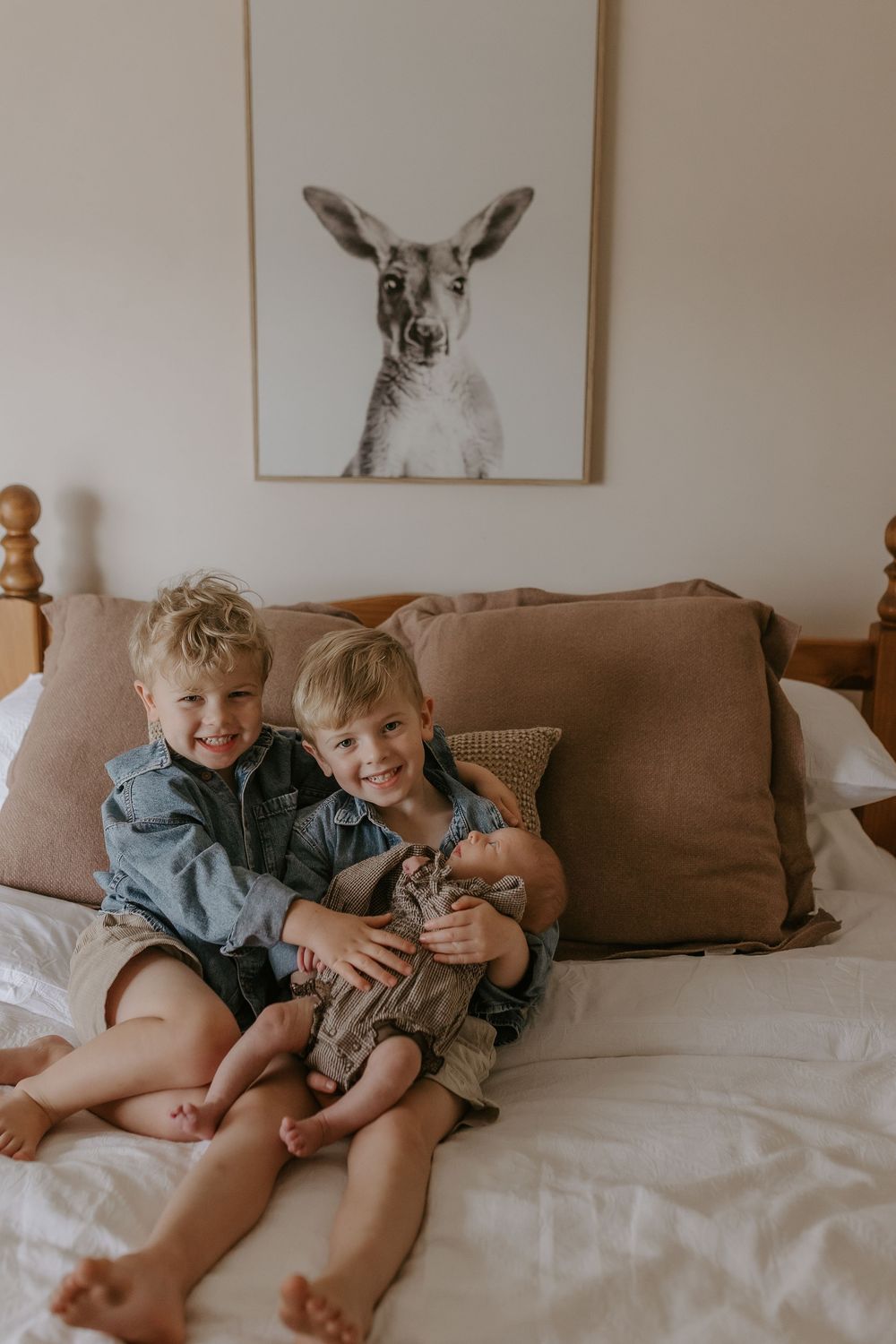 Two smiling young boys sitting on a bed holding a newborn baby with a kangaroo wall print behind them.