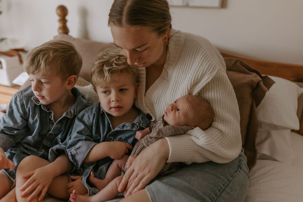 A mother sits on a bed holding a newborn while two young toddler boys cuddle close beside her.